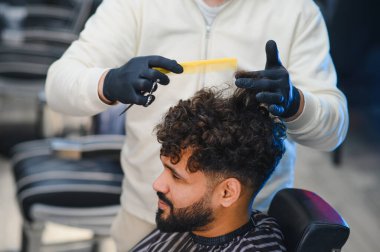 Profesyonel berber bir adamın kıvırcık saç stilini makas ve tarak ile buduyor, tımar ve güzelliğe odaklanıyor.