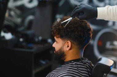Berber, bir adamın kıvırcık saçlarını kesmek için makas kullanarak yeni bir görünüm yaratıyor. Bakım ve stile odaklanma