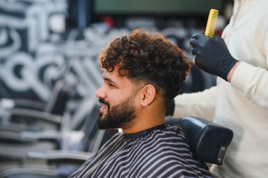 Gülümseyen adam modern berberdeki eldivenli berberden şık bir tarak ve sakal kesimi alıyor, profesyonel tımar hizmetinden zevk alıyor.