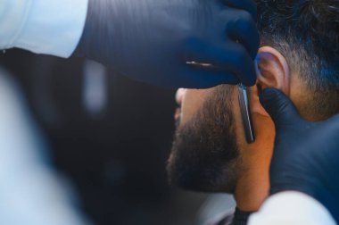 Bir adamın sakalını ve favorilerini titizlikle traş etmek ve kesmek için keskin bir jilet kullanan eldivenli eller.