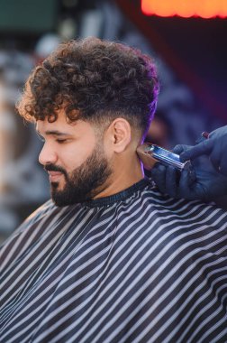 Kıvırcık saçlı bir adam eldiven takan bir berberden taze, soluk bir saç tıraşı oluyor, şık bir bakım ve tımara odaklanıyor.