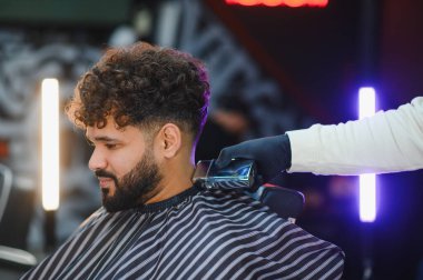 Kıvırcık saçlı ve sakallı bir adam elektrikli makasla berberberden saç tıraşı oluyor.