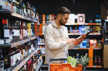 Süpermarket koridorunda şarap şişesi etiketini inceleyen bir adam alışveriş arabasının yanında durup ürünleri seçmeye odaklanmış.