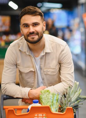 Genç adam gülümsüyor, marketin koridorunda sağlıklı yiyeceklerle dolu bir alışveriş arabasına yaslanıyor. Kameraya bakmak, tüketici yaşam tarzı kavramı