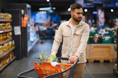 Parlak bir süpermarkette alışveriş yapan sakallı bir adam, dolu bir arabayı iterken düşünceli bir şekilde bakkaliye ve taze ürünleri seçiyor.