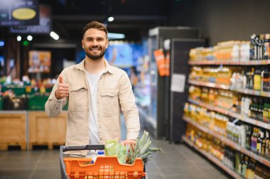 Süpermarkette bir araba dolusu erzağı iten, gülümseyen ve başparmağını kaldıran bir adam.