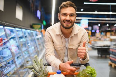 Süpermarkette genç bir adam taze ürünlerle dolu bir arabayı itiyor, gülümsüyor ve harika bir bakkal seçimi için başparmağını kaldırıyor.