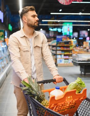Taze ürün ve içeceklerle dolu bir market arabasını iten adam, parlak ışıklı süpermarket koridorlarında günlük ihtiyaçlarını seçiyor.