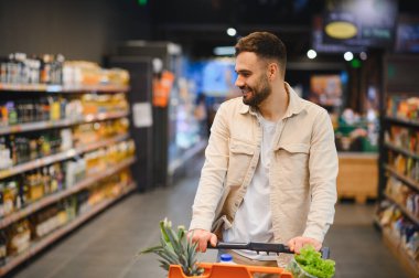 Gülümseyen adam ürünler buluyor ve kalabalık bir market koridorunda alışveriş yaparken sağlıklı yiyecekler seçiyor.