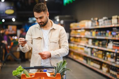 Alışveriş arabasındaki yiyecekleri bulanık bir süpermarket geçmişiyle karşılaştıran adam alışveriş listesi ve bütçeye odaklanıyor.