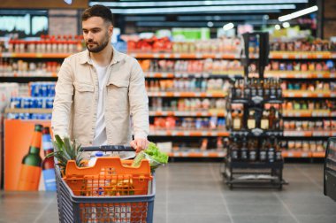 Genç adam, marketin yoğun koridorunda içi taze ürünlerle dolu bir alışveriş arabasını itiyor.