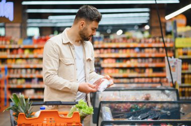 Modern bir süpermarkette içi yiyecek dolu bir alışveriş arabasını iterken paketlenmiş ete bakan bir adam.