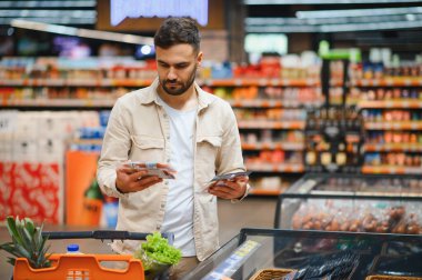 Elinde iki paket yiyecek olan bir adam marketin koridorunda alışveriş arabasıyla seçenekler arasında karar veriyor.