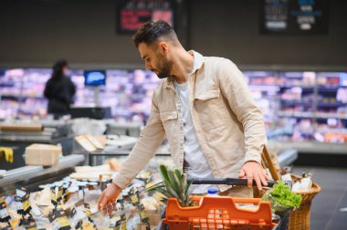 Genç bir adam süpermarkette soğutuculu bir vitrinden peynir seçiyor, market arabasıyla alışveriş yapıyor.