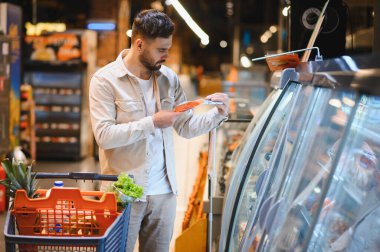 Market alışverişi yapan, süpermarkette soğutulmuş bir kasede paketlenmiş et ürününü inceleyen bir adam.