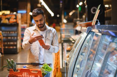 Genç adam markette alışveriş arabasını iterken paketlenmiş somon filetosunun etiketini okuyor.