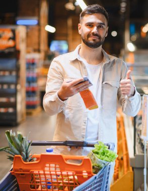 Telefonu ve kredi kartını elinde tutan mutlu bir adam, taze yiyecek dolu bir alışveriş arabasını iterken başparmağını kaldırıyor.