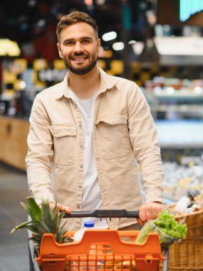 Genç adam, marketten yiyecek alma tecrübesinin tadını çıkararak, içi taze ürünlerle dolu bir alışveriş arabasını itiyor.