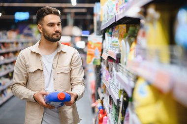 Elinde bir şişe deterjan tutan bir adam, marketin ev eşyaları bölümündeki raflardan ürünleri seçiyor.