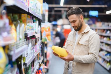 Sarı bir şişe tutan, ürün etiketini okuyan ve perakende mağazasının ev eşyaları bölümünde seçenekleri karşılaştıran bir adam.