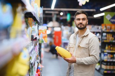 Elinde sarı temizleme deterjanı olan bir adam marketin koridorunda dikiliyor. Ev malzemesi seçen tüketici
