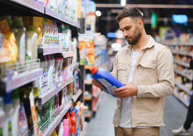 Çamaşır deterjanı şişesini inceleyen bir adam markette raflardaki temizlik malzemeleri arasından seçim yapıyor.