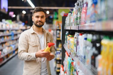 Süpermarkette elinde sprey şişeyle dolaşan bir adam, raflardaki temizlik ürünlerini karşılaştırırken hangisini alacağına karar veriyor.