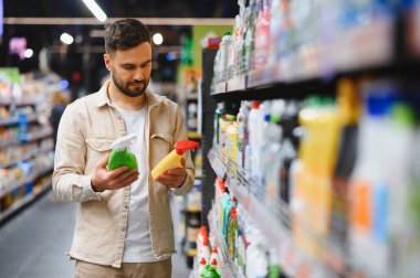 Alışveriş yapan kişi, süpermarket koridorunda iki şişe temizlik ürününü karşılaştırıyor ve seçenekler arasında seçim yapıyor.