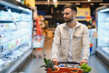 Genç bir adam taze ürünlerle dolu bir alışveriş arabasını itiyor, marketteki soğutulmuş raflardaki ürünlere bakıyor.