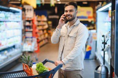 Gülümseyen bir adam alışveriş arabasını iterken market koridorunda telefonla konuşuyor.