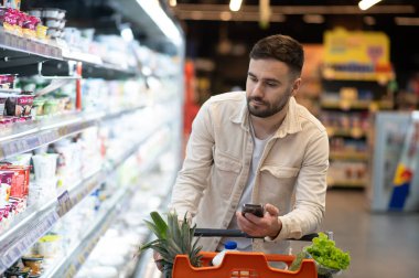 Bir markette ürün rafının yanında duran genç adam elinde telefonla alışveriş arabasındaki eşyalara bakıyor.