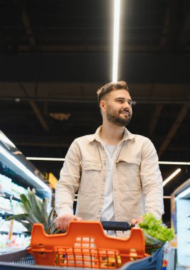 Market koridorunda içi taze meyve ve sebze dolu alışveriş arabasını iten adam.