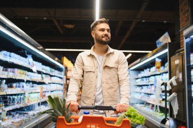 Taze ürünlerle dolu bir alışveriş arabasını iten adam, parlak bir bakkal koridorunda sağlıklı yiyecekler seçiyor.