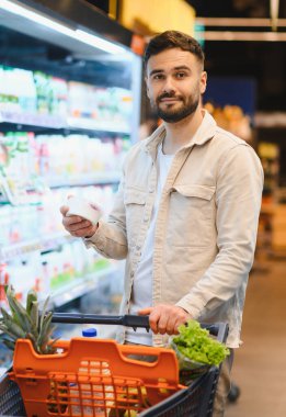 Taze ürünlerle dolu bir alışveriş arabasını iten adam marketin soğutulmuş bölümünden bir ürün seçiyor.