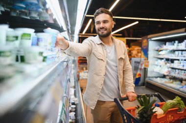 Dondurulmuş süt ürünleri reyonunda duran genç bir adam, bir bakkal arabasını iterken yoğurt ürününe uzanıyor.
