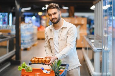 Genç adam bir alışveriş arabası itiyor, süpermarket koridorunda taze gıda ürünleri arıyor. Sağlıklı yaşam tarzı ve günlük alışveriş kavramı