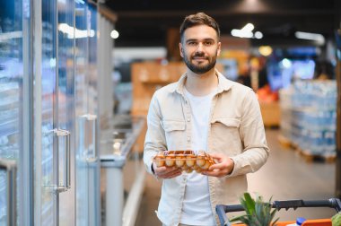 Genç adam bakkal alışverişi yapıyor, elinde yumurta kutusu tutuyor ve modern bir süpermarkette alışveriş arabasını itiyor.