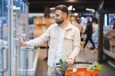 Genç bir adam market alışverişi yapıyor, buzdolabının kapısını açıyor. Taze yiyeceklerle dolu bir arabayı itiyor.
