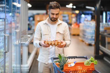 Bir markette içi taze meyve ve diğer şeylerle dolu bir alışveriş arabasını iterken elinde bir kutu yumurta tutan bir adam.