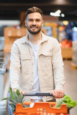 Genç adam gülümserken içi taze ürünlerle dolu bir alışveriş arabasını itiyor. Modern bir süpermarket koridorunda alışveriş yapıyor.