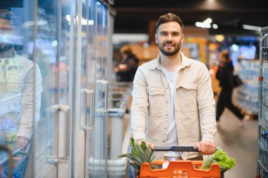 Gülümseyen, içi taze ürünlerle dolu bir alışveriş arabasını parlak bir marketten iten genç adam.