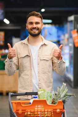 Süpermarkette alışveriş arabasıyla duran gülümseyen bir adam olumlu onay hareketi gösteriyor.