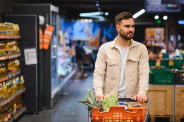 Genç bir adam, parlak bir süpermarket koridorunda içi taze yiyeceklerle dolu bir alışveriş arabasını itip ürün arıyor.