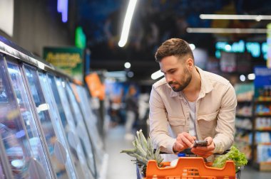Akıllı telefondan alışveriş listesini kontrol eden bir adam, modern bir markette dondurulmuş gıda reyonunun yakınındaki taze ürünlerle dolu alışveriş arabalarını itiyor.