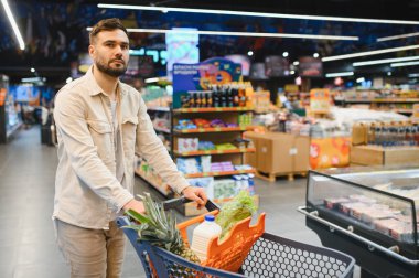 Genç adam taze ürünler, süt ve konserve ürünlerle dolu bir alışveriş arabasını satıyor, perakende mağazasından alışveriş yapıyor.