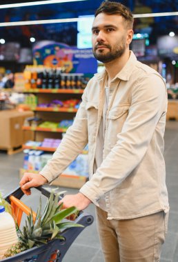 Müşteri bakkal reyonunda ürün seçiyor, yiyecek dolu bir alışveriş arabasını itiyor.