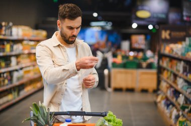 Süpermarket koridorunda duran bir adam, elinde uzun bir makbuzla, gıda fiyatları ve bütçe artışı konusunda kafası karışık ve endişeli hissediyor.