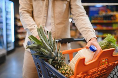 Bir adamın elleri marketin koridorunda içi taze ürün, süt ve diğer şeylerle dolu bir alışveriş arabasını itiyor.