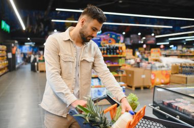 Genç adam alışveriş arabasını itiyor, taze ürünleri ve süt ürünlerini parlak bir süpermarket koridorunda seçiyor.