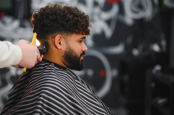 Kıvırcık saçlı ve sakallı bir adam modern bir kuafördeki bir berberden elektrikli makasıyla yeni bir saç kesimi alıyor.